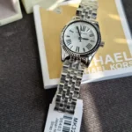 Silver Michael Kors wristwatch with a Roman numeral dial resting on a gold Michael Kors box, metal link bracelet visible.