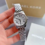 Silver Michael Kors wristwatch with white dial and Roman numerals held in hand, metal link bracelet visible in background.