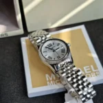 Silver Michael Kors wristwatch with Roman numeral dial resting on a yellow-beige box.