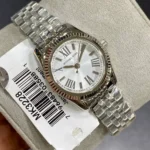 Silver Michael Kors wristwatch with Roman numerals, white dial, and brushed stainless steel bracelet shown on a display cushion with a barcode tag nearby.