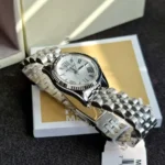 Silver stainless steel wristwatch with a fluted bezel and Roman numeral dial resting on a card and its box.