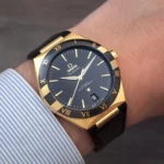 omega-constellation-co-axial-master-chronometer-41-mm-leather-strap-automatic_5 Gold-tone Omega Constellation wristwatch on a striped shirt cuff, black dial with gold Roman numerals.