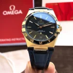 omega-constellation-co-axial-master-chronometer-41-mm-leather-strap-automatic_6 Gold Omega Constellation wristwatch with black dial, gold Roman numerals on bezel, and blue leather strap; date window at 6 o'clock.