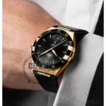 omega-constellation-co-axial-master-chronometer-41-mm-leather-strap-automatic_7 Gold-tone and black two-tone wristwatch on a man's wrist with a black leather strap and cuffed shirt sleeve.