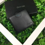 Black Calvin Klein wallet resting on a black box against a backdrop of green plants; metal brand logo visible on the wallet corner.