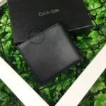 Black leather wallet resting on a Calvin Klein box on a green leafy background.