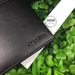 Close-up of a black leather Calvin Klein wallet with the brand name on the corner, resting on a white card among green leaves.