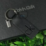 Black leather Calvin Klein keychain with a metal ring and small rectangular tag on a dark textured surface.