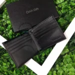 Open black Calvin Klein leather wallet showing card slots on a green leafy background with the CK box nearby.