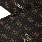 Close-up of black textured material with brown repeating monogram pattern and a white GUESS logo triangle visible at the bottom edge, and a zipper seam crossing the image.