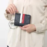 original-tommy-hilfiger-wallet-women-navy-blue-genuine-leather_11 Small black textured wallet with white and red vertical stripes and a gold logo, held by a person wearing a beige blouse/coat.