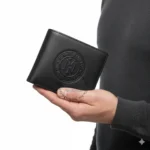Black leather bi-fold wallet with a circular embossed logo being held in a hand.