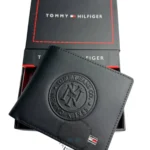 Black leather Tommy Hilfiger wallet with embossed circular logo, shown with black box and red stitching detail in the background