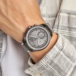 tommy_hilfiger_max_1791975_1 Metal wristwatch with grey dial on a wrist, metal band, wearing a striped jacket sleeve visible in the background.