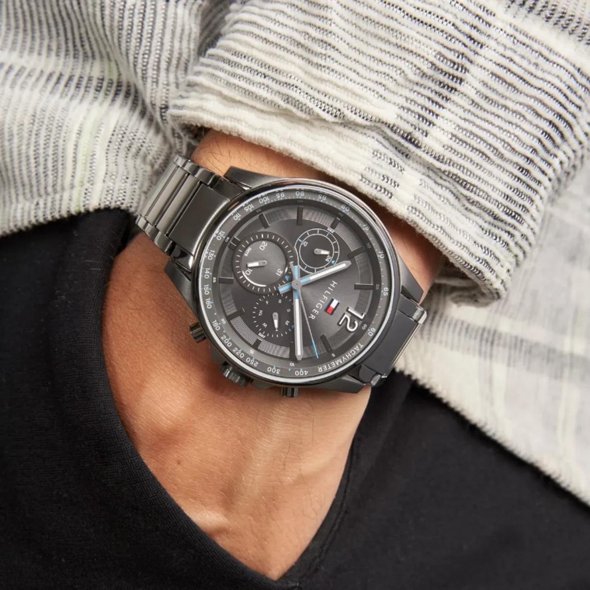 Close-up of a black metal chronograph watch worn on a wrist over a striped knit sleeve.
