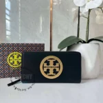 Black zip wallet with a large gold circular emblem, placed on a white surface beside a patterned box and a white orchid planter.