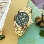 Gold Michael Kors wristwatch with a black dial and three subdials on a colorful surface.