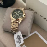 Gold Michael Kors chronograph wristwatch with a green dial resting on a beige cushion, with a box and price tag nearby.