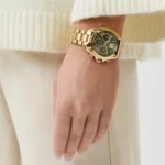 Gold wristwatch with black dial worn on a left wrist, over a cream knit sweater sleeve portrait.