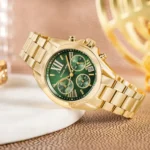 Gold-toned Michael Kors chronograph watch with a green dial and date window, displayed on a textured surface.
