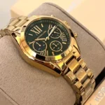 Gold-tone Michael Kors chronograph watch with a dark green dial, Roman numerals, three subdials, and a date display, resting on a gray cushion.