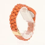 Orange metal link watch on a clear display stand, shown from the side with a visible clasp edge.