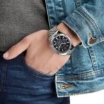 mk8280-michael-kors-watch-men-blue-dial-metal-stainless-steel-silver-strap-quartz-battery-analog-chronograph-lexington_107 Wrist with a silver metal watch and black dial, worn beside a denim jacket sleeve and gray sweater.