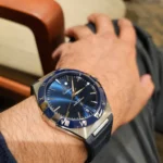 Wristwatch with blue dial and Roman numeral bezel worn on a hairy forearm, showing a stainless steel case and blue strap.