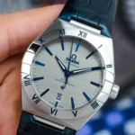 Omega Constellation wristwatch with silver dial, blue hour markers, and blue hands on a dark strap.
