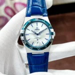 Omega Constellation watch with white dial, blue Roman-numeral bezel, and blue leather strap, held in gloved hand.