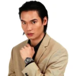 Man in a tan blazer and black shirt posing with his left hand near his collar, wearing a silver wristwatch against a white background.