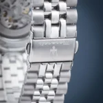 Close-up of a polished stainless steel watch bracelet with a deployment clasp, engraved branding on the clasp and a blurred watch movement in the background.