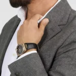 r8851153001-original-maserati-watch-men-black-dial-leather-strap_9 Man in a gray check blazer adjusts his white shirt collar, wearing a gold-tone watch with a black leather strap on his wrist.