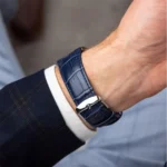 Blue crocodile-embossed leather watch strap on a man's wrist, silver buckle peeking from a dark suit sleeve.