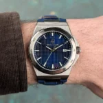 Stainless steel wristwatch with a blue textured dial and a date window at 3 o'clock, worn on a wrist with a brown sleeve.