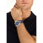 Man wearing a blue-faced wristwatch with a stainless steel case and blue strap, his hand resting on his neck in a black polo shirt.