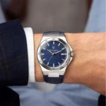 Silver stainless steel wristwatch with a blue textured dial on a man's wrist, dressed in a dark suit. (Date window at 3 o'clock)
