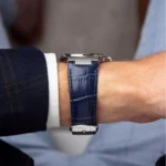 Close-up of a wrist wearing a silver watch with a blue crocodile-pattern leather strap, partially tucked under a dark suit sleeve with a white shirt cuff visible.