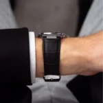 Wrist wearing a black leather strap watch with a metal case, visible cuff of a white shirt and dark suit sleeve in a formal setting