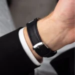 Close-up of a left wrist wearing a black leather strap watch with a silver buckle, white shirt cuff, and dark suit sleeve.