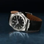 A stainless steel wristwatch with a black textured dial, white indices, and a leather strap, showing the date window at 3 o'clock.