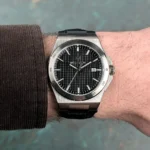 Stainless steel wristwatch with a black textured dial, silver hour markers, and a date window at 3 o'clock on a brown-sleeved wrist