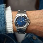 Stainless steel wristwatch with a blue textured dial on a man's wrist, denim sleeve visible on the left.