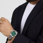 Man wearing a silver metal wristwatch with a green dial on his left wrist, paired with a black blazer and white shirt.