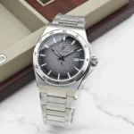 Silver stainless steel wristwatch with a textured dark gray dial and a date window at 3 o'clock on a marble surface.