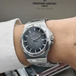 Stainless steel wristwatch with a dark textured dial and date window on the right, worn on a cuffed wrist.