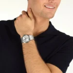 Man in a black polo shirt shows off a silver stainless steel wristwatch on his left wrist while touching his neck and smiling.