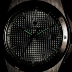 Close-up of a stainless steel wristwatch with green luminous hands and markers on a textured black dial; date window at 3 o'clock.
