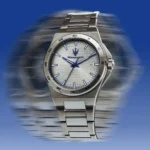 Maserati stainless steel wristwatch with a textured light dial, blue hands and a date window at 3 o'clock, against a blurred blue backdrop