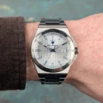 Stainless steel wristwatch with a textured silver dial, blue hour markers, and a date window at 3, worn on a forearm with a brown cuffed sleeve.